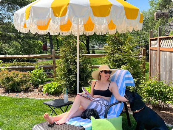 Chic backyard setup featuring a vintage-inspired Pagoda Cut patio umbrella in yellow and white Sunbrella fabric with elegant fringe detailing. A stylish woman lounges under the shade, enjoying a book with her two dogs, creating a relaxed and sophisticated outdoor retreat.
