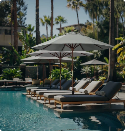 Lifestyle image showcasing the Grove Series Patio Umbrellas by California Umbrella, elegantly shading lounge chairs by a luxurious poolside surrounded by lush greenery, emphasizing their timeless design and durability for upscale hospitality and resort settings.