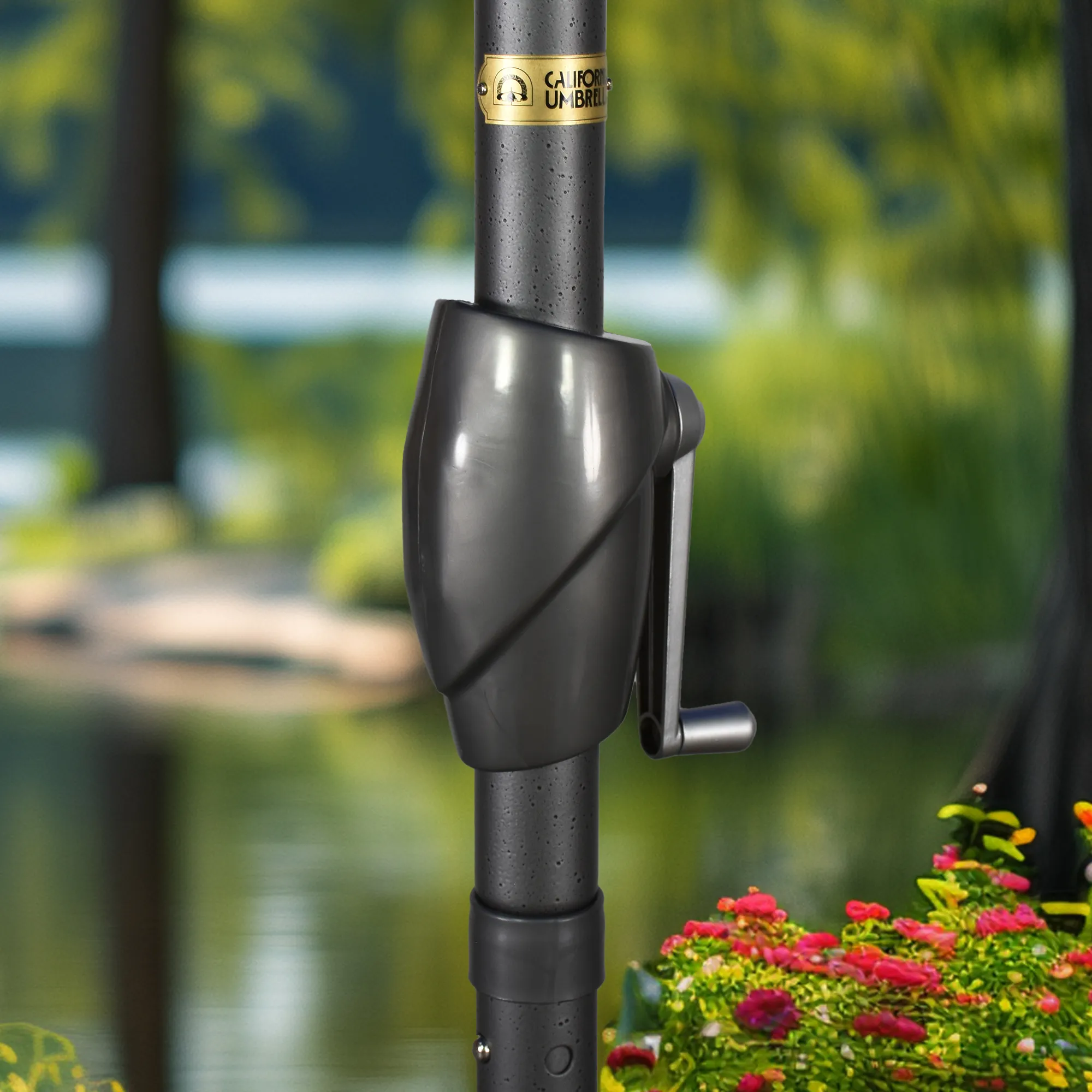 Close-up image of the easy crank lift mechanism on the Pacific Trail Series Patio Umbrella by California Umbrella. This detailed view highlights the ergonomic crank handle set against a serene garden backdrop, illustrating the simplicity and functionality of the umbrella's operation. The crank lift, featured in a sleek grey finish, allows for effortless opening and closing, ensuring convenient shade adjustment that enhances the user experience in any outdoor setting.